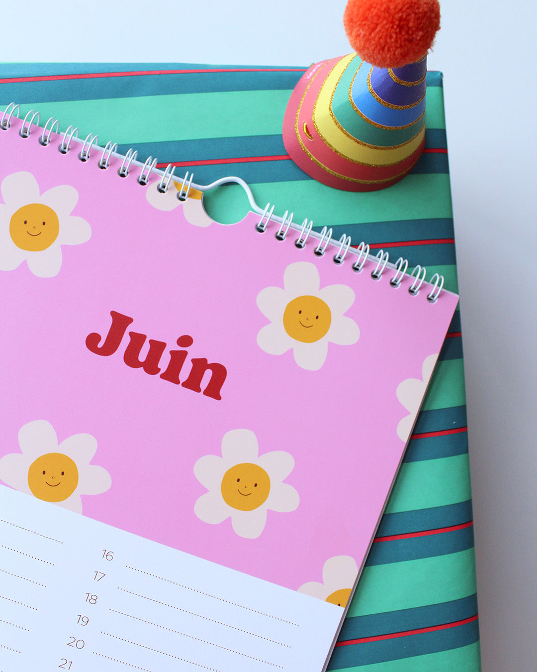 Un vibrant calendrier des anniversaires Ma Petite Vie est ouvert au mois de juin, affichant "Juin" en rouge sur une page rose avec des fleurs blanches souriantes. Il est posé sur une boîte cadeau rayée, à côté d'un chapeau de fête assorti, surmonté d'un pompon rouge.