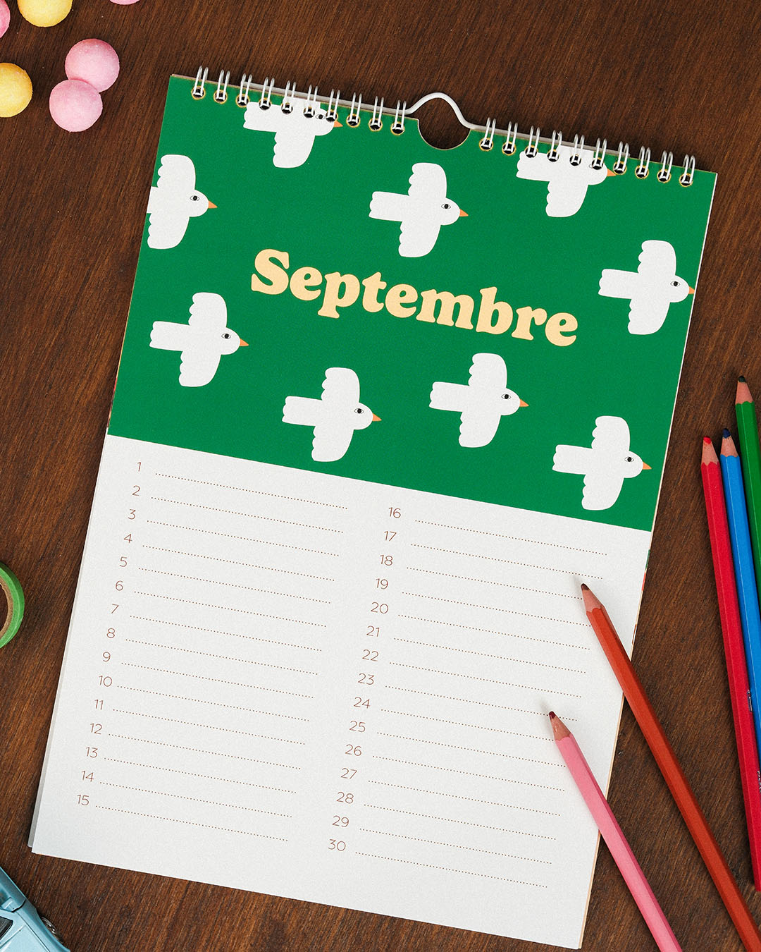 Le "Calendrier des anniversaires" de Ma Petite Vie est ouvert à la page "Septembre" verte et blanche avec des illustrations d'oiseaux, posée sur une table en bois avec des crayons de couleur et des bonbons - un cadeau familial idéal.