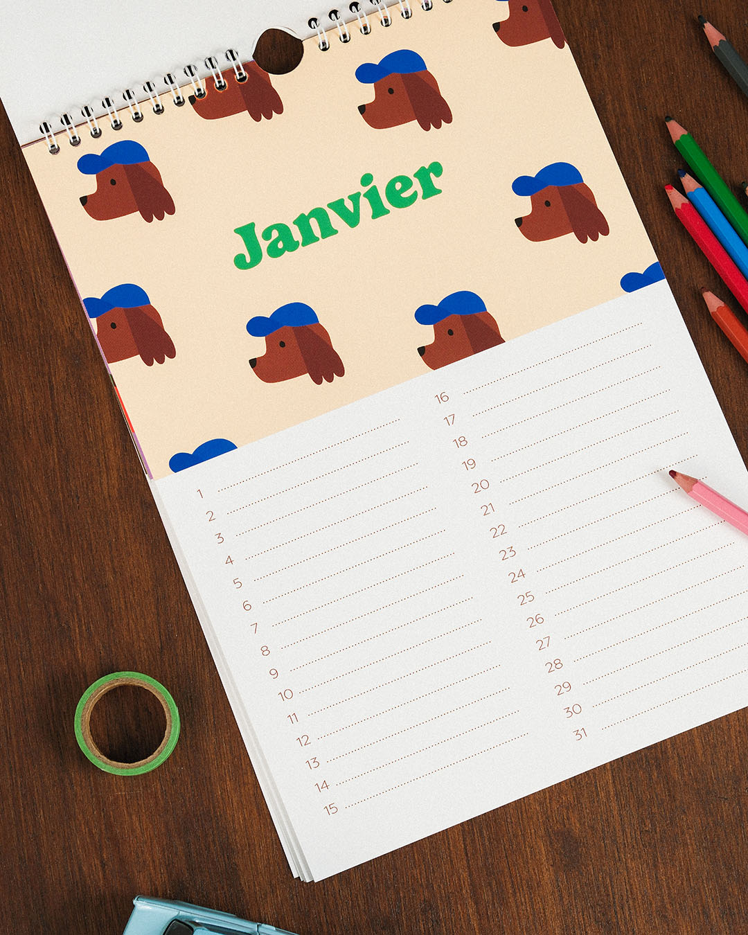 Le Calendrier des anniversaires de Ma Petite Vie s'ouvre sur "Janvier" avec un chien de dessin animé coiffé d'une casquette bleue, des crayons de couleur, du ruban adhésif vert et un taille-crayon bleu - idéal pour un cadeau familial.